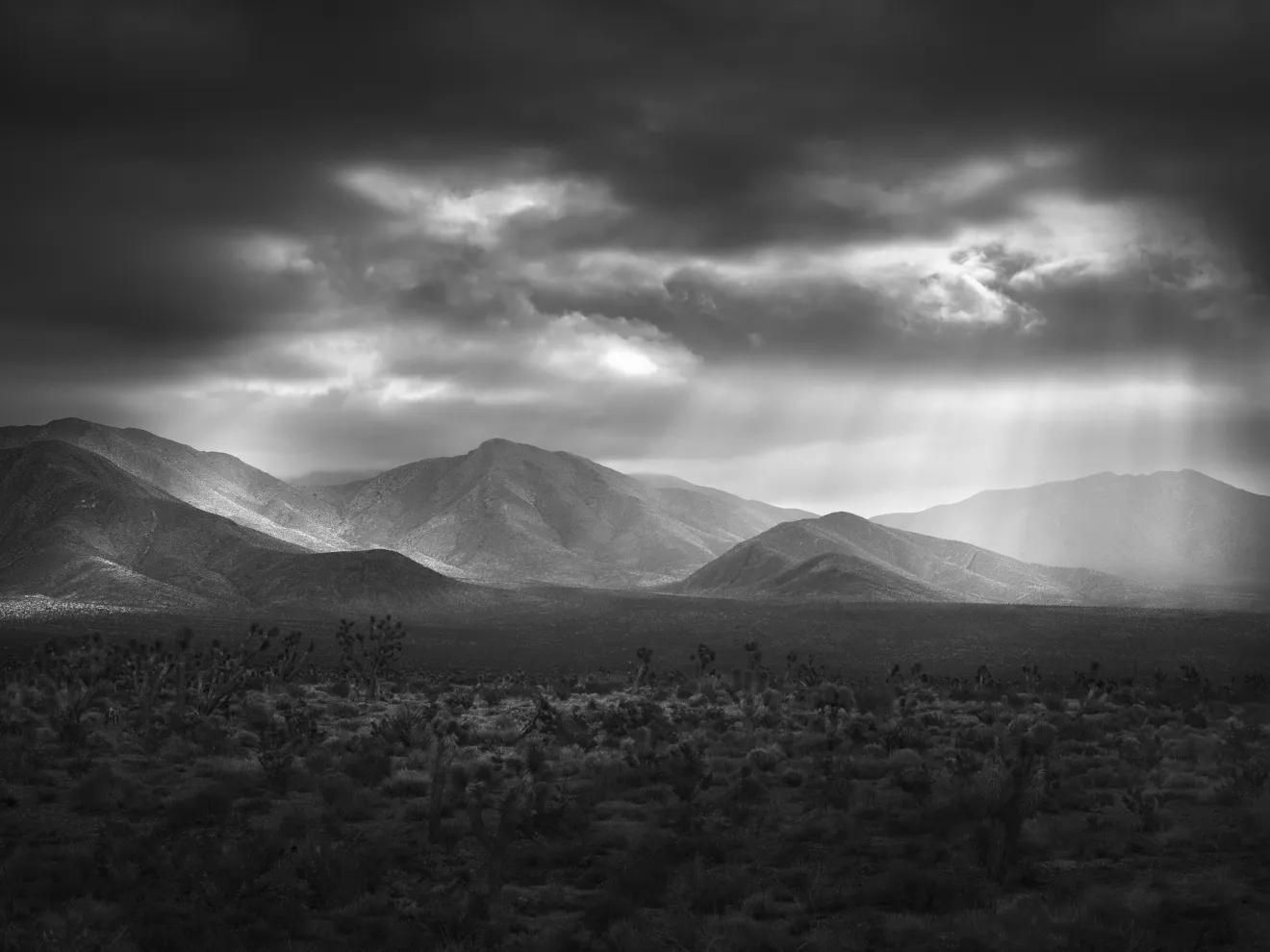 Black and white landscape of mountains under a cloudy sky with sunlight streaming through.