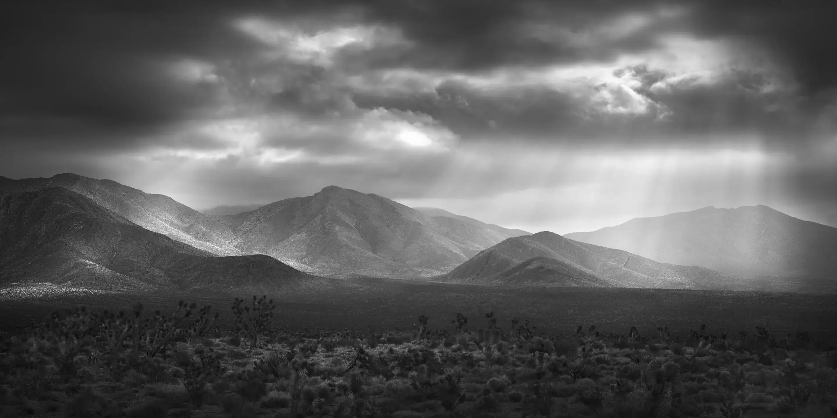 Sunlight breaks through clouds, illuminating a range of distant mountains in a dramatic black-and-white scene.
