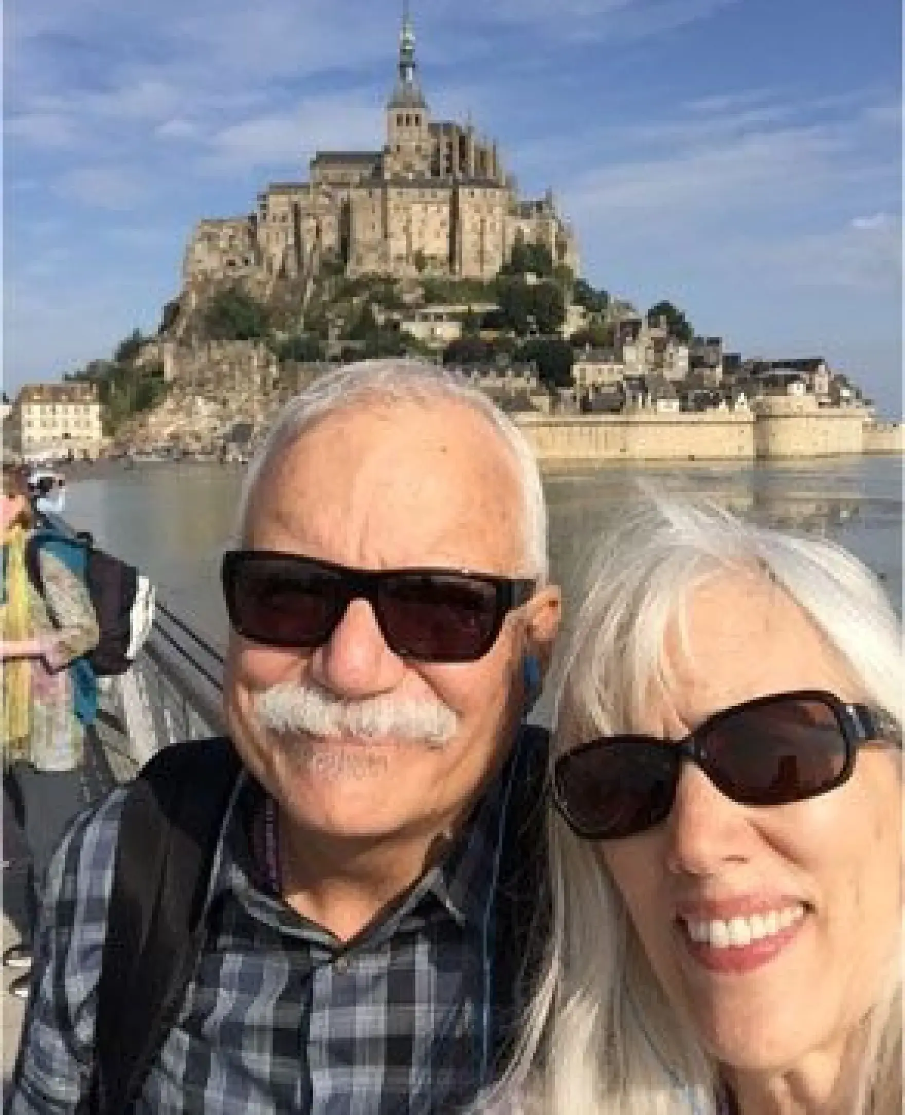 An older couple wearing sunglasses smiles while posing in front of Mont Saint-Michel.