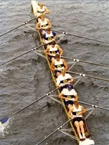 Eight people rowing in a long boat on the water, viewed from above.